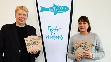 Ein Mann und eine Frau halten ein Kochbuch für Fischgerichte in die Kamera | © Dörte Behrmann