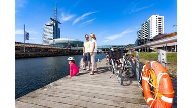 Zwei Erwachsene und ein Kind stehen auf einem Steg am Wasser, daneben stehe ihr Fahrräder. | © Helmut Gross / Erlebnis Bremerhaven GmbH