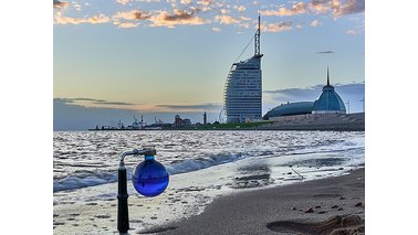 Ein mit blau gefärbtem Wasser gefüllter Rundkolben auf Stativ in Richtung des Klimahauses. | © Torsten Tingler