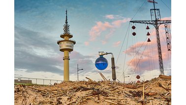 Ein mit blau gefärbtem Wasser gefüllter Rundkolben auf Stativ in Richtung des Radarturms mit Wasserstandsanzeiger. | © Torsten Tingler