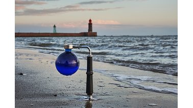 Ein mit blau gefärbtem Wasser gefüllter Rundkolben auf Stativ mit Blick auf Weser und Mole . | © Torsten Tingler