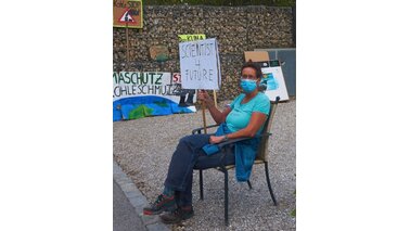 Eine Frau sitzt auf einem Stuhl und hält ein beschriebenes Schild hoch. | © Dr. Manuela Troschke