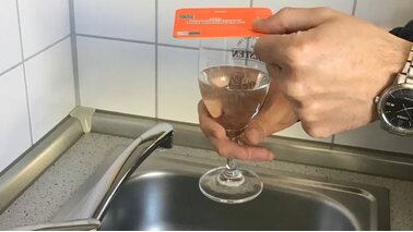 Eine Hand hält auf ein mit Wasser befülltes Glas einen orangenen Bierdeckel.  | © Sven Passarge