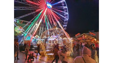 Eine Fotoaufnahme auf einem Weihnachtsmarkt am Abend mit beleuchteten Riesenrad und Lichtelementen.  | © Tanja Mehl