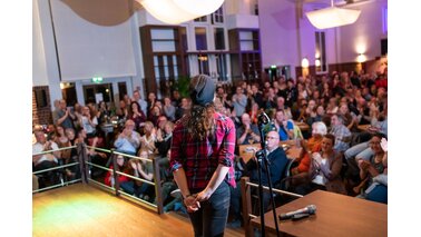 Eine Dame steht auf einer Bühne vor Publikum, welches ihr aufmerksam bei Ihren Vortrag zuhört.