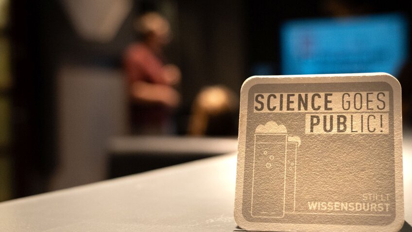 Das ist ein grauer Bierdeckel mit der Aufschrift "Science Goes Public!" 