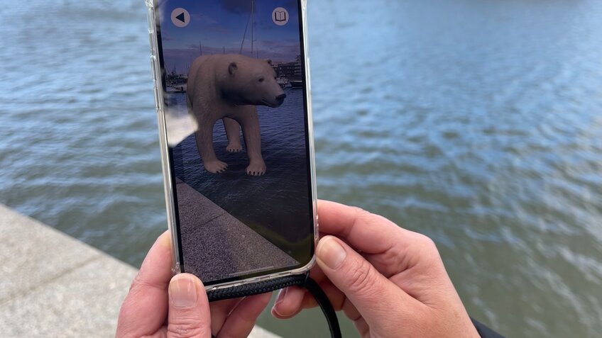 Eine Person Hält ein Handy auf der die Bremerhaven Guide App vor dem Zoo Am Meer gezeigt wird mit einem Eisbären auf dem Bildschirm